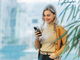Porträt einer Frau im Business-Casual-Look, die lächelnd auf ihr Handy schaut. Warme Erdtöne und heller, moderner Hintergrund.