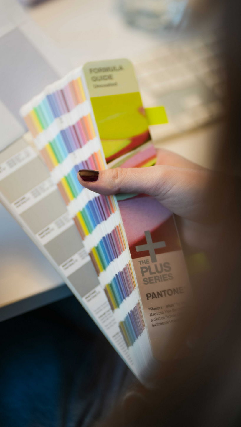 Nahaufnahme aus der Perspektive über die Schulter einer Designerin, die einen aufgeschlagenen Pantone-Farbfächer in der Hand hält, um Farben abzugleichen