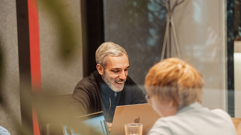 Ein freundlich lächelnder Mann sitzt im Besprechungsraum und unterhält sich konzentriert mit einer Kollegin bei der gemeinsamen Projektarbeit am Laptop