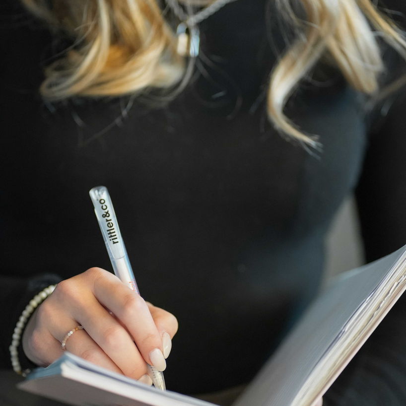 Eine Person mit blonden Haaren hält ein offenes Notizbuch mit Spiralbindung und schreibt mit einem Stift, auf dem das Logo „rittler&co“ aufgedruckt ist