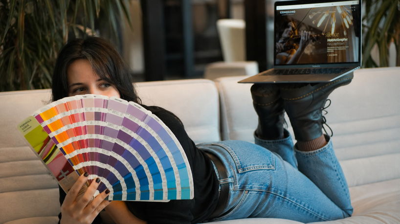 Eine Frau liegt auf einem hellen Sofa und hält einen aufgefächerten, bunten Pantone-Farbfächer vor ihr Gesicht. Auf ihren angewinkelten Beinen balanciert sie ein geöffnetes Laptop, das eine moderne Website zeigt