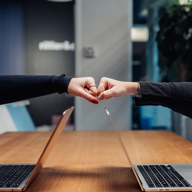 Zwei Personen sitzen sich an einem hölzernen Schreibtisch mit aufgeklappten Laptops gegenüber und formen mit ihren Händen gemeinsam ein Herz – Symbol für Teamwork und ein positives Arbeitsklima.