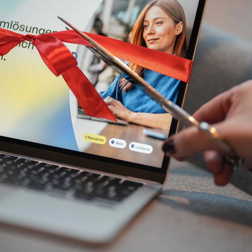 Nahaufnahme eines Laptops, auf dessen Bildschirm eine neue Website zu sehen ist. Um den Bildschirm ist eine rote Geschenkschleife gebunden, die im Vordergrund symbolisch mit einer Schere durchschnitten wird