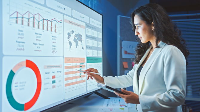 Eine Frau in Businesskleidung steht vor einem großen Bildschirm, der detaillierte Diagramme, Karten und Analysedaten zeigt. Sie verwendet einen Stift, um auf spezifische Datenpunkte zu zeigen, während sie ein Tablet in der anderen Hand hält.