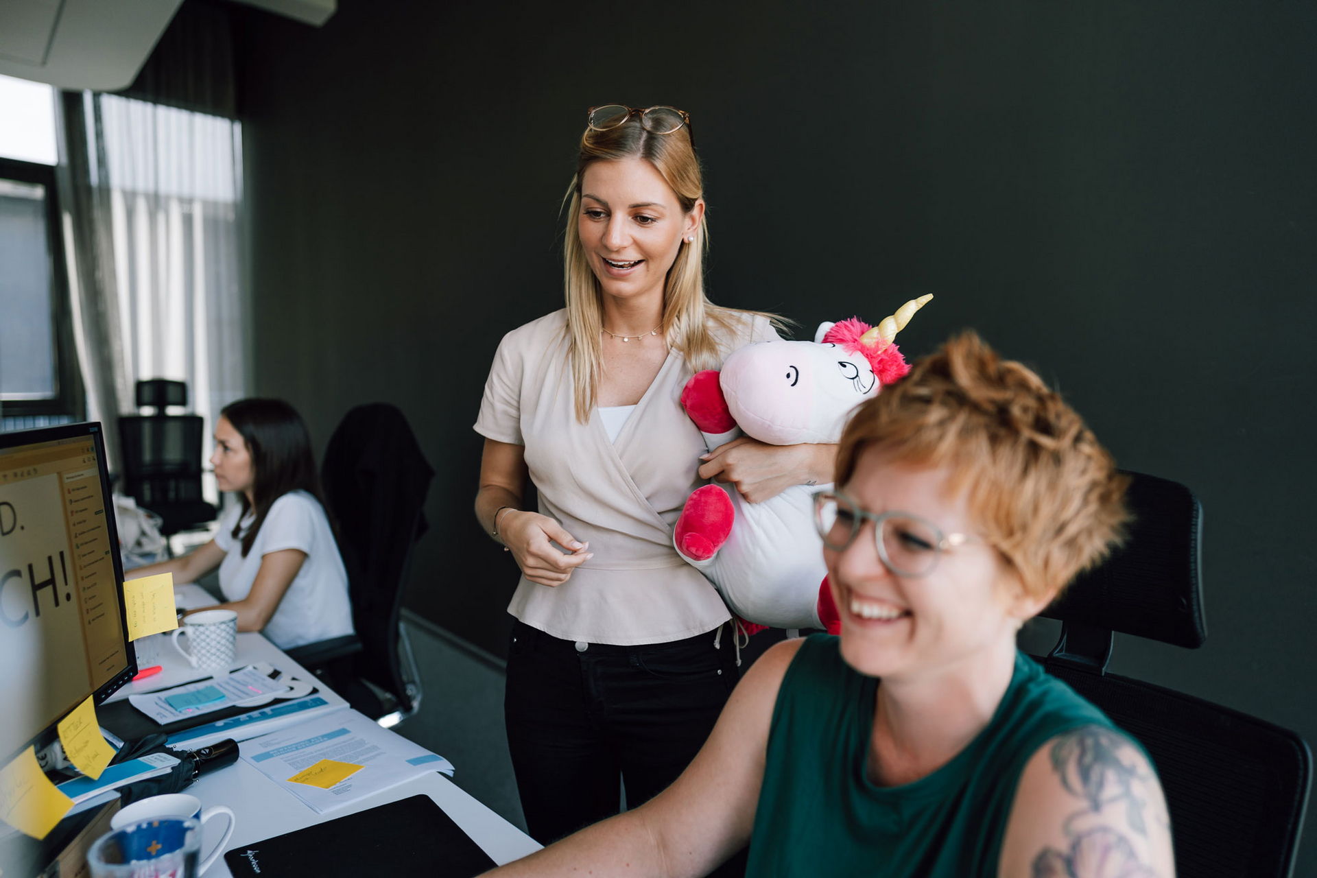 Frau mit Einhorn Plüschtier sieht Designerin bei der Arbeit über die Schulter