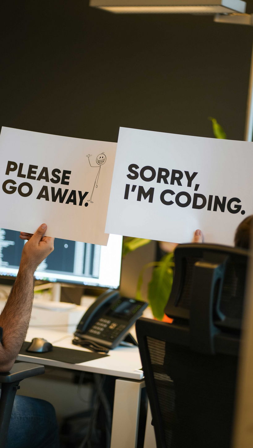Ein Foto eines Büroschreibtisches, an dem eine Person humorvoll Schilder hochhält mit den Texten „PLEASE GO AWAY“ und „SORRY, I'M CODING.