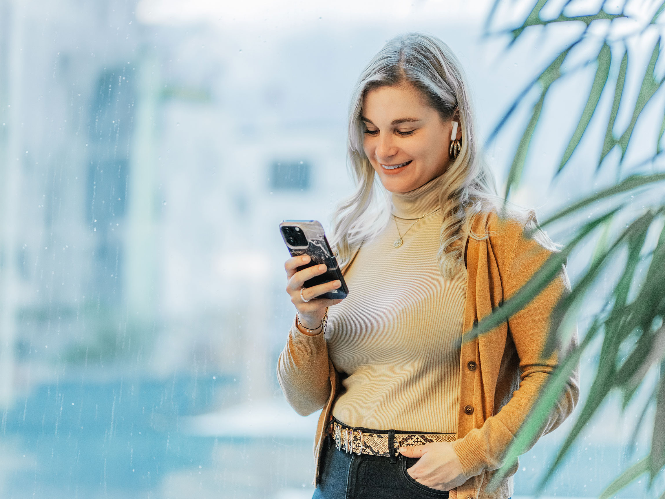 Porträt einer Frau im Business-Casual-Look, die lächelnd auf ihr Handy schaut. Warme Erdtöne und heller, moderner Hintergrund.